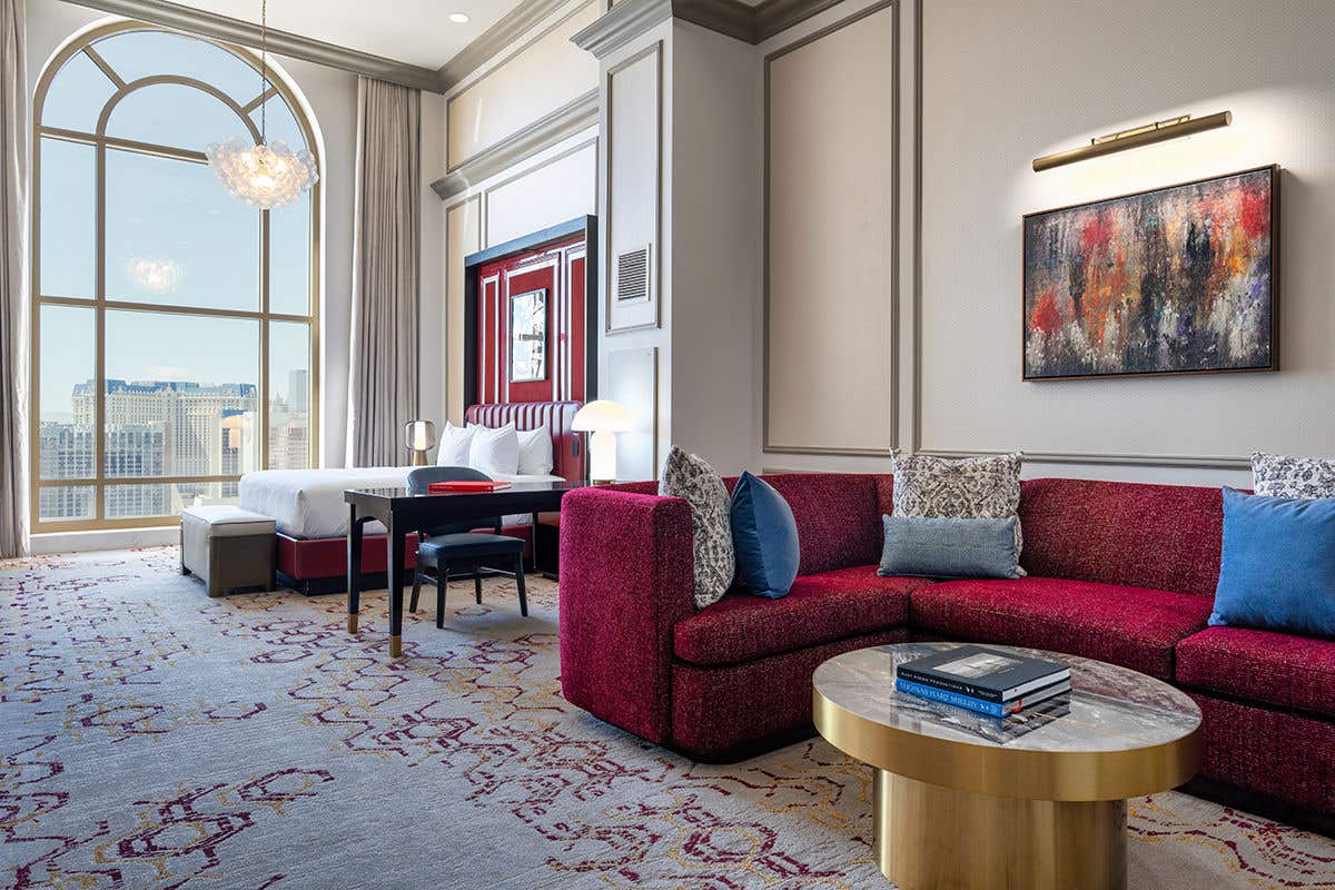 A luxurious hotel suite with a king bed, a red sectional sofa, and a large arched window overlooking the city.