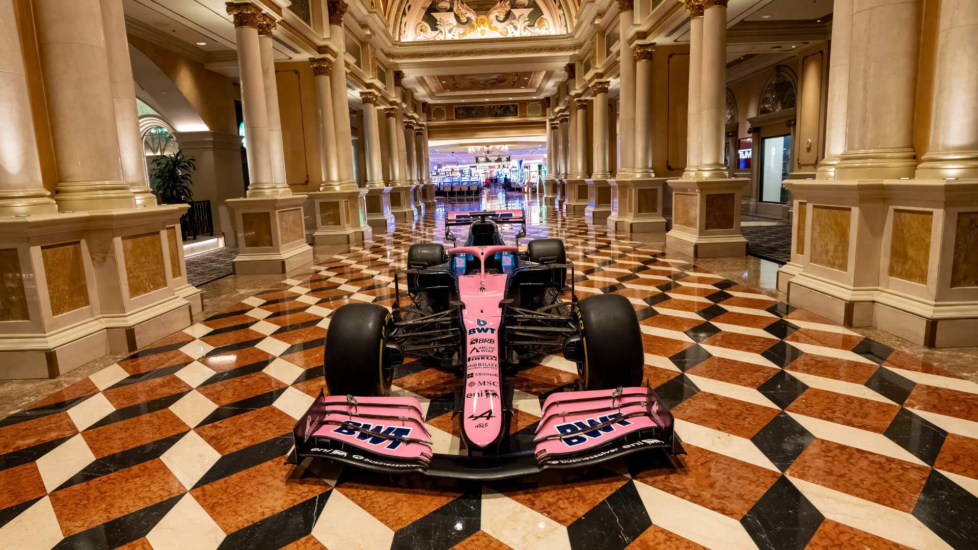 Front view of a pink and blue Formula 1 race car displayed indoors on a patterned marble floor inside an ornate hallway with columns and decorative ceilings at The Venetian Resort Las Vegas.