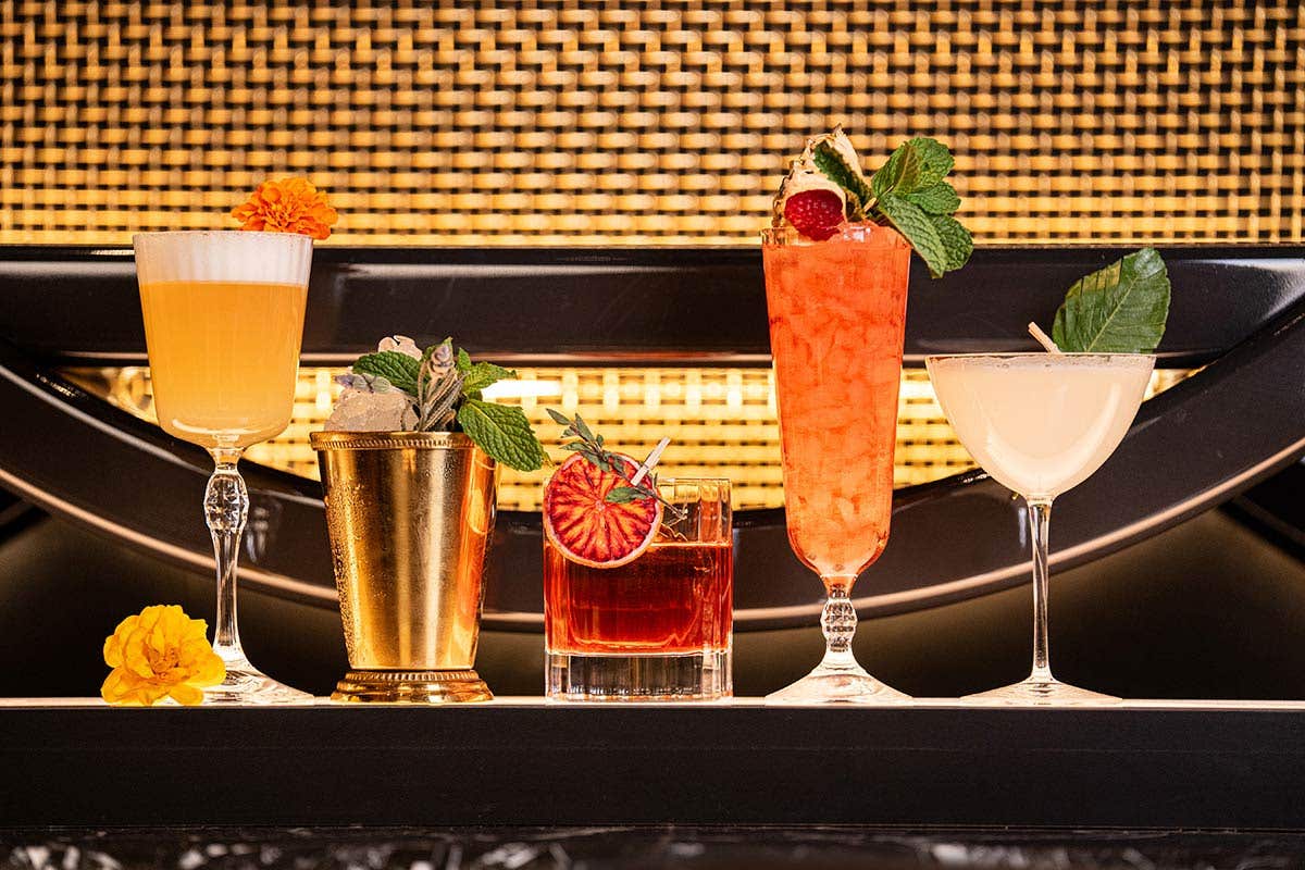 Five cocktails in various glasses on a bar top against a golden patterned background