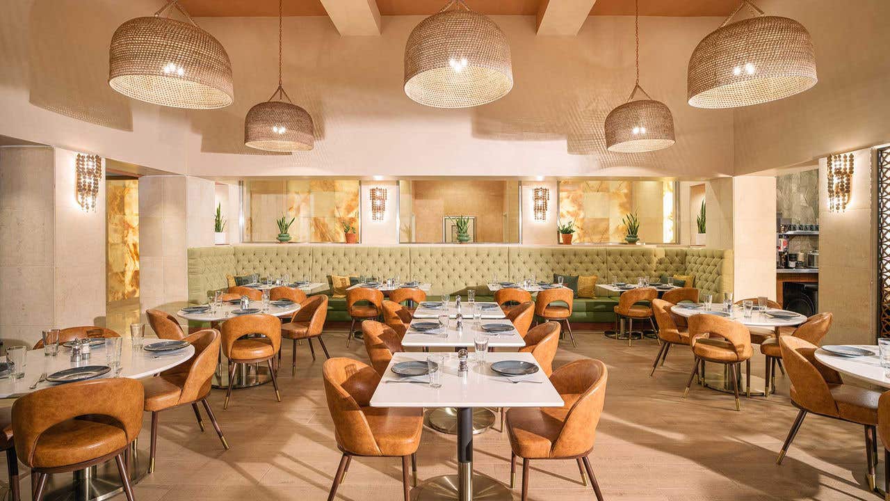 Modern restaurant interior with warm neutral tones, featuring multiple square tables set with plates and glassware, surrounded by tan leather chairs, a long upholstered banquette along the back wall, and large woven pendant lights hanging from the ceiling.
