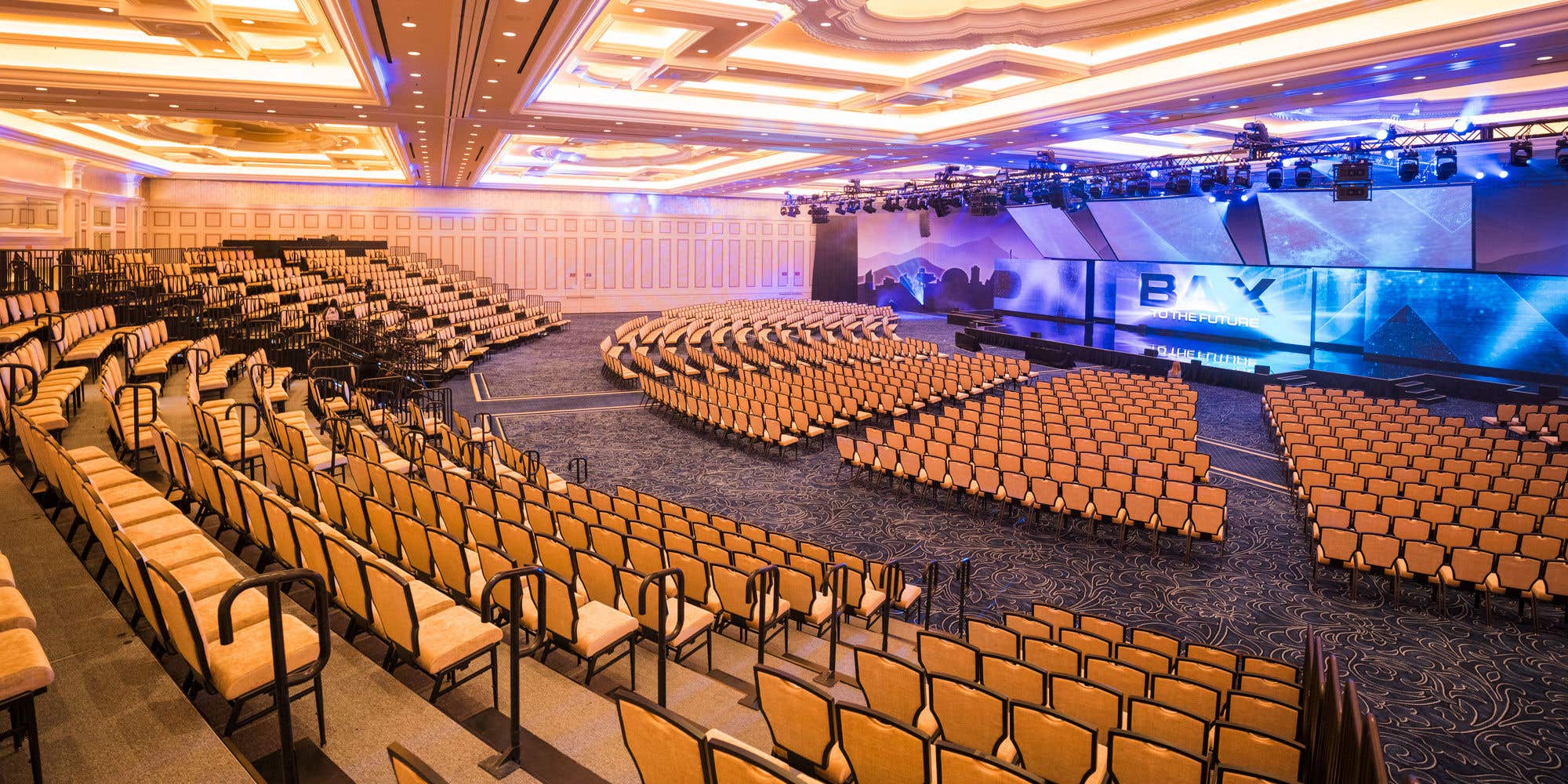 A large, theater-style conference room with tiered seating faces a modern stage with a large screen, all under an ornate, illuminated ceiling.