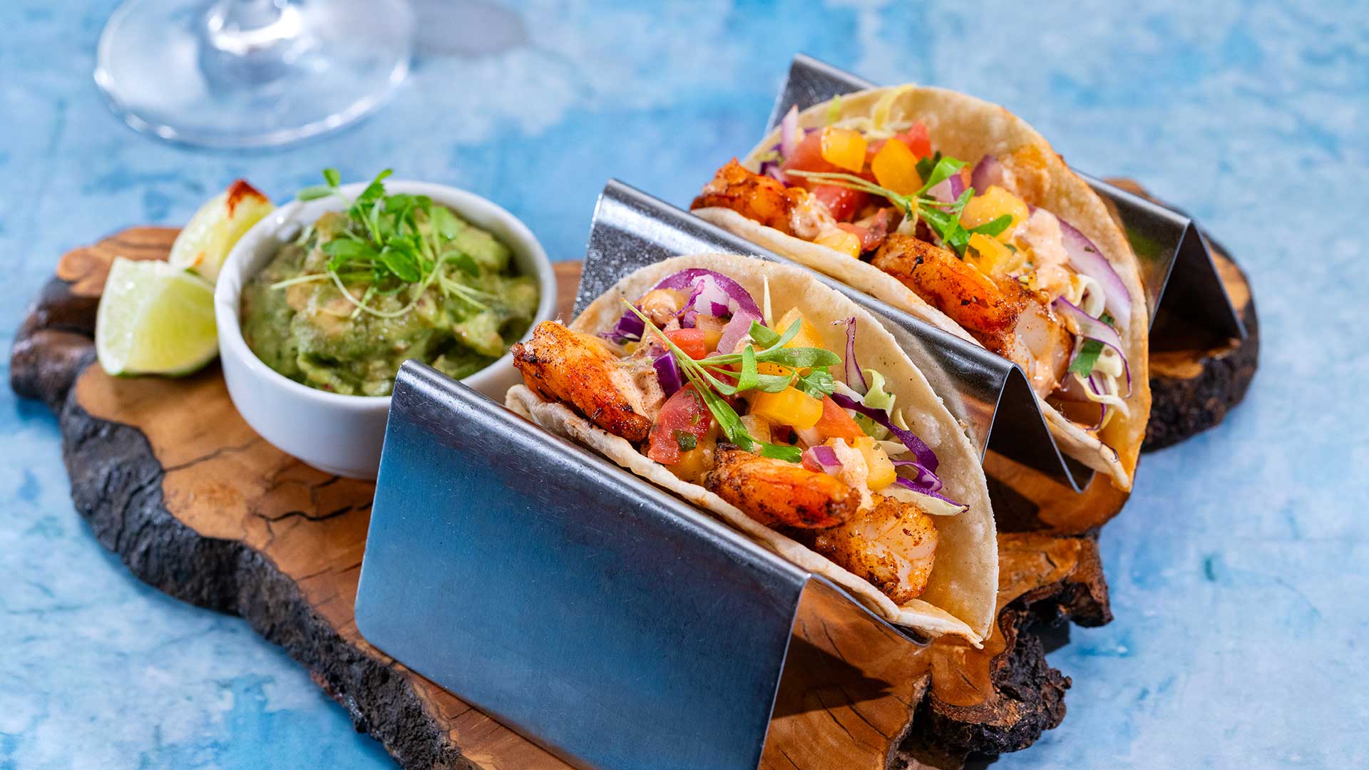 Two soft tacos filled with grilled shrimp, diced peppers, shredded cabbage, and herbs are held upright in metal taco stands on a rustic wooden board, with a small bowl of guacamole and a lime wedge on a blue textured surface.