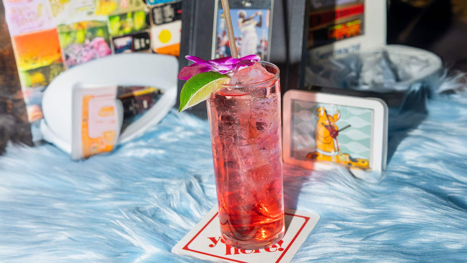 A tall Collins glass with a red drink and hibiscus and lime garnish, set on a blue fur rug. ‘90s memorabilia is visible behind.