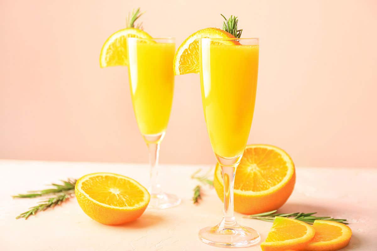 Two tall champagne flutes filled with a bright orange cocktail, garnished with orange slices and sprigs of rosemary, set on a light surface with halved oranges and slices arranged nearby against a soft peach background.
