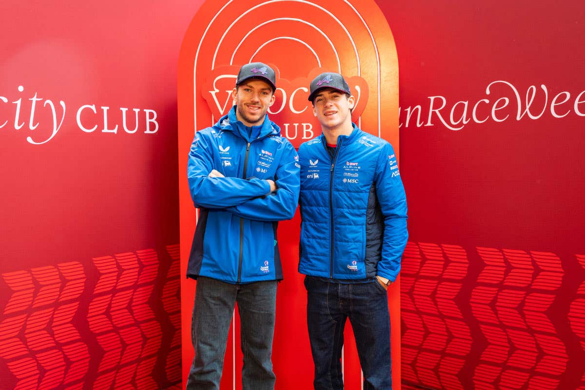 Two people wearing blue Alpine team jackets standing side by side in front of a red backdrop with “Velocity Club” and event branding visible.