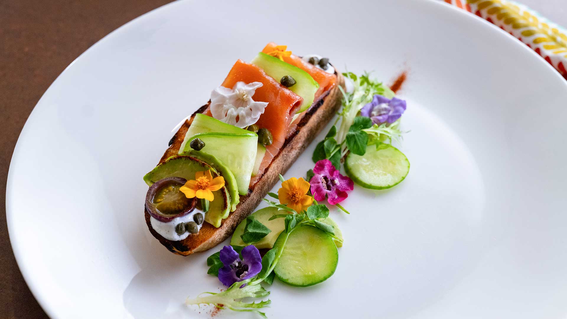 Toast topped with salmon, avocado slices, capers, and edible flowers, plated with cucumber and greens.